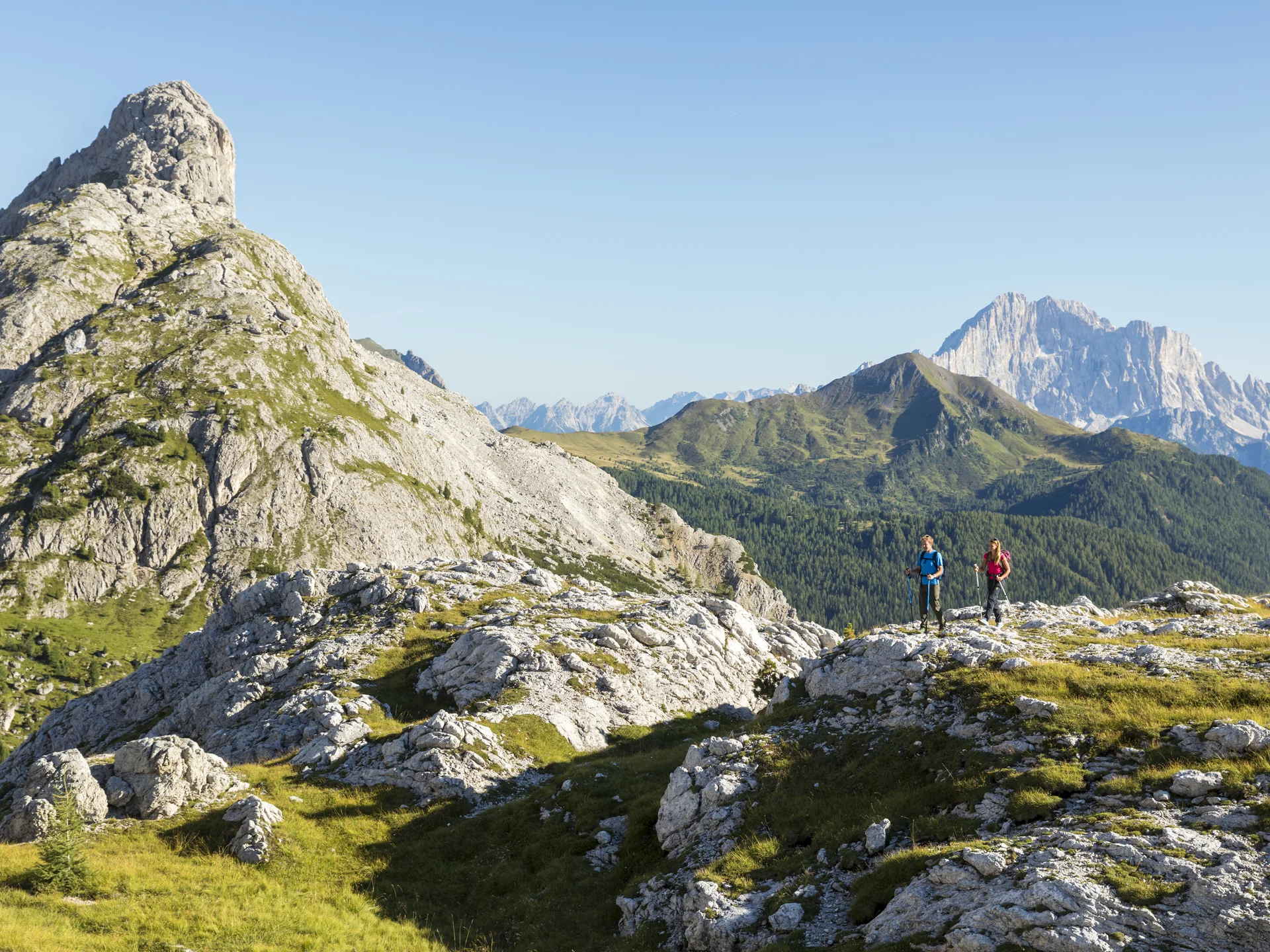Two hikers on rocky trail in green mountain landscape under clear sky