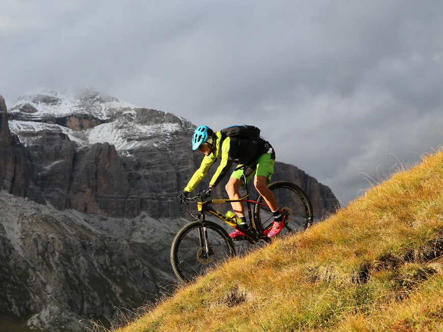 Cyclist wearing blue helmet rides down grassy slope with rocky mountains in background