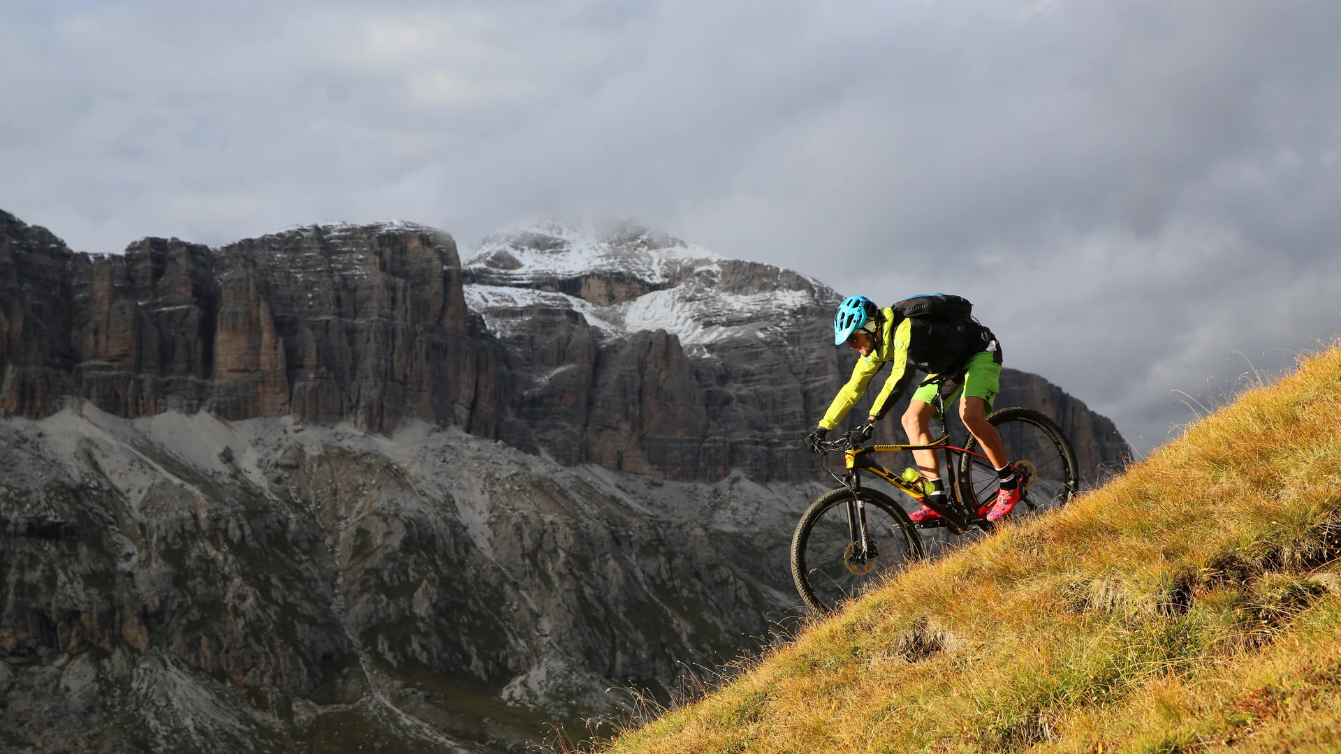 Bilder aus Arabba des Chalet Barbara Radfahrer mit blauem Helm fährt einen grasbewachsenen Hang vor felsigen Bergen hinunter
