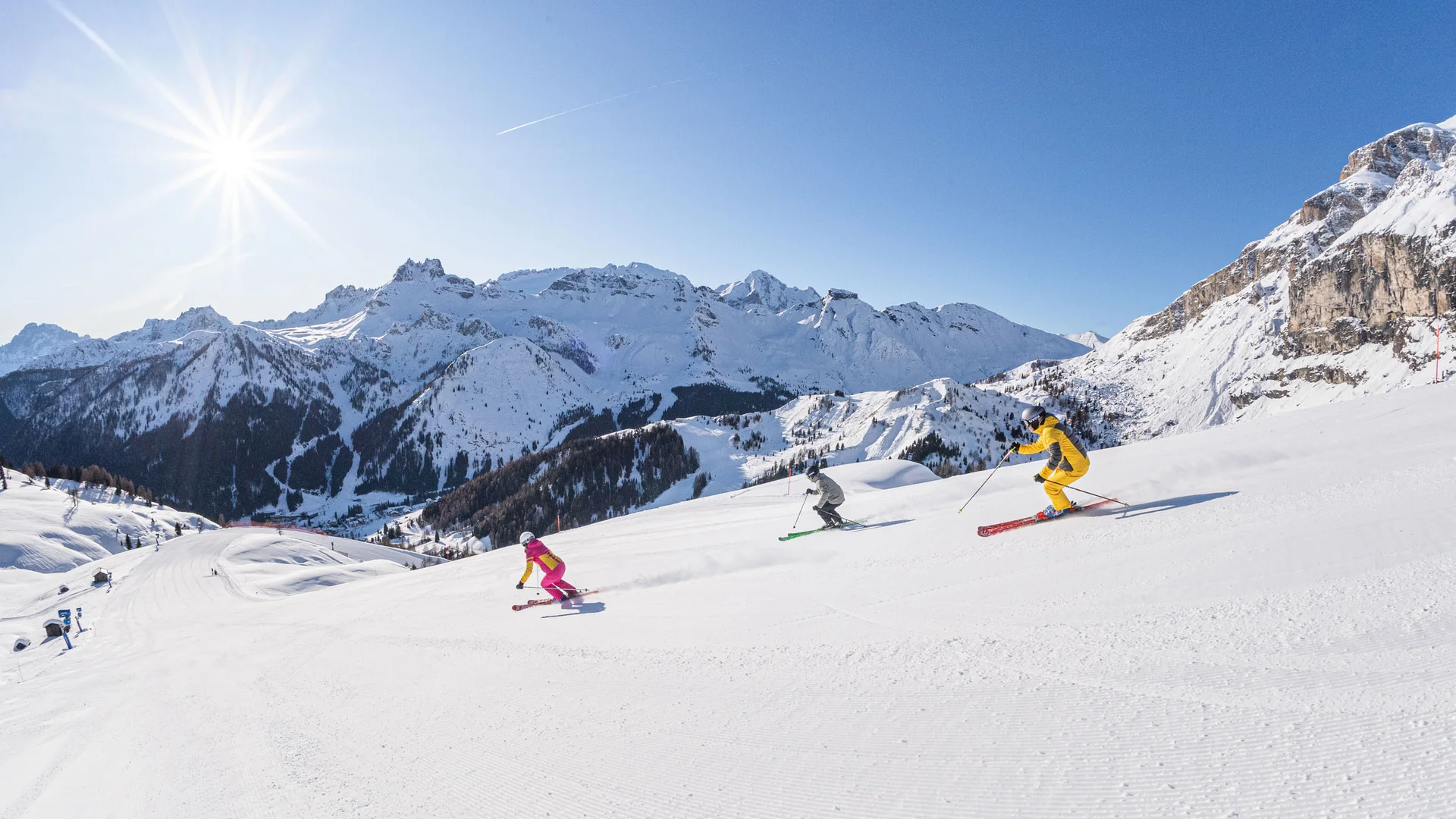 Bilder aus Arabba des Chalet Barbara Drei Skifahrer in bunter Kleidung fahren eine verschneite Piste mit Bergen im Hintergrund hinab