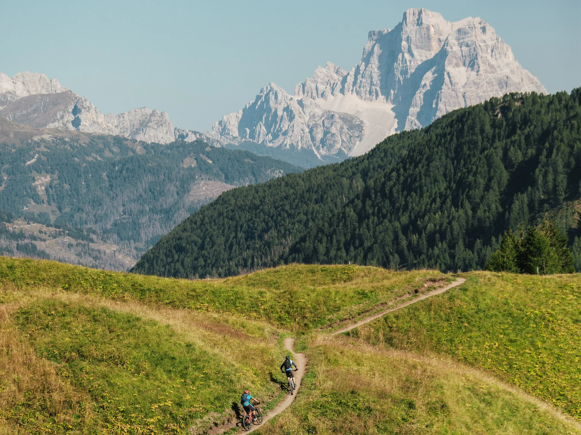 Two cyclists on a trail in grassy hills with rocky mountains in the background