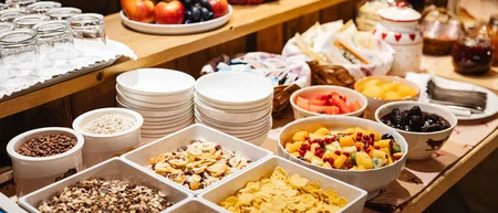 Frühstücksbuffet mit Müsli, frischem Obst und gestapeltem Geschirr auf einem Holztisch