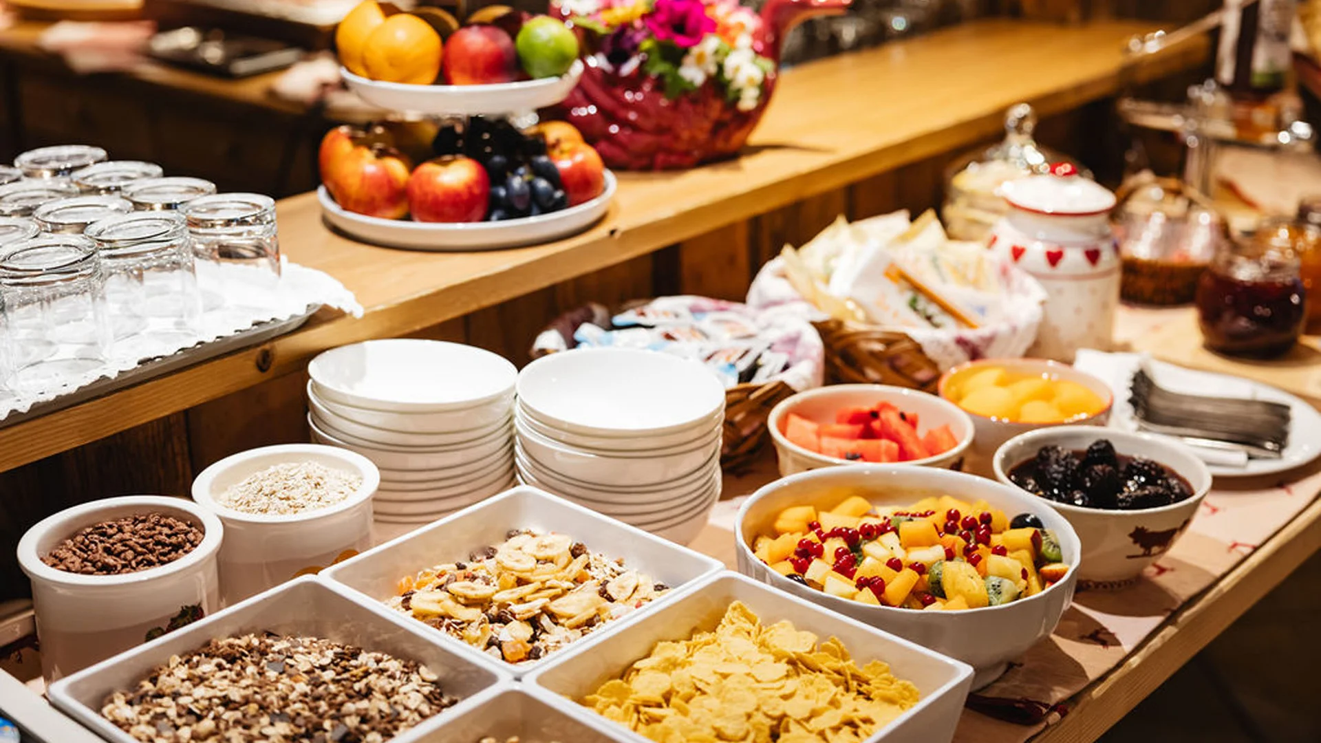 Bilder aus Arabba des Chalet Barbara Frühstücksbuffet mit Müsli, frischem Obst und gestapeltem Geschirr auf einem Holztisch
