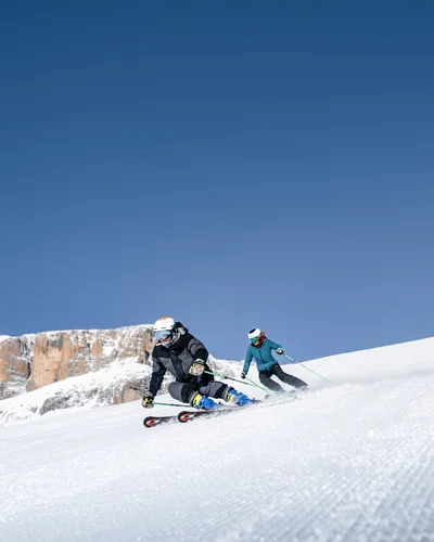 Two skiers skiing down a snowy slope with mountains in the background
