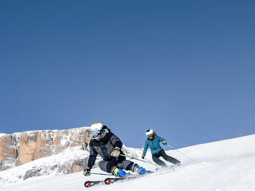 Two skiers skiing down a snowy slope with mountains in the background