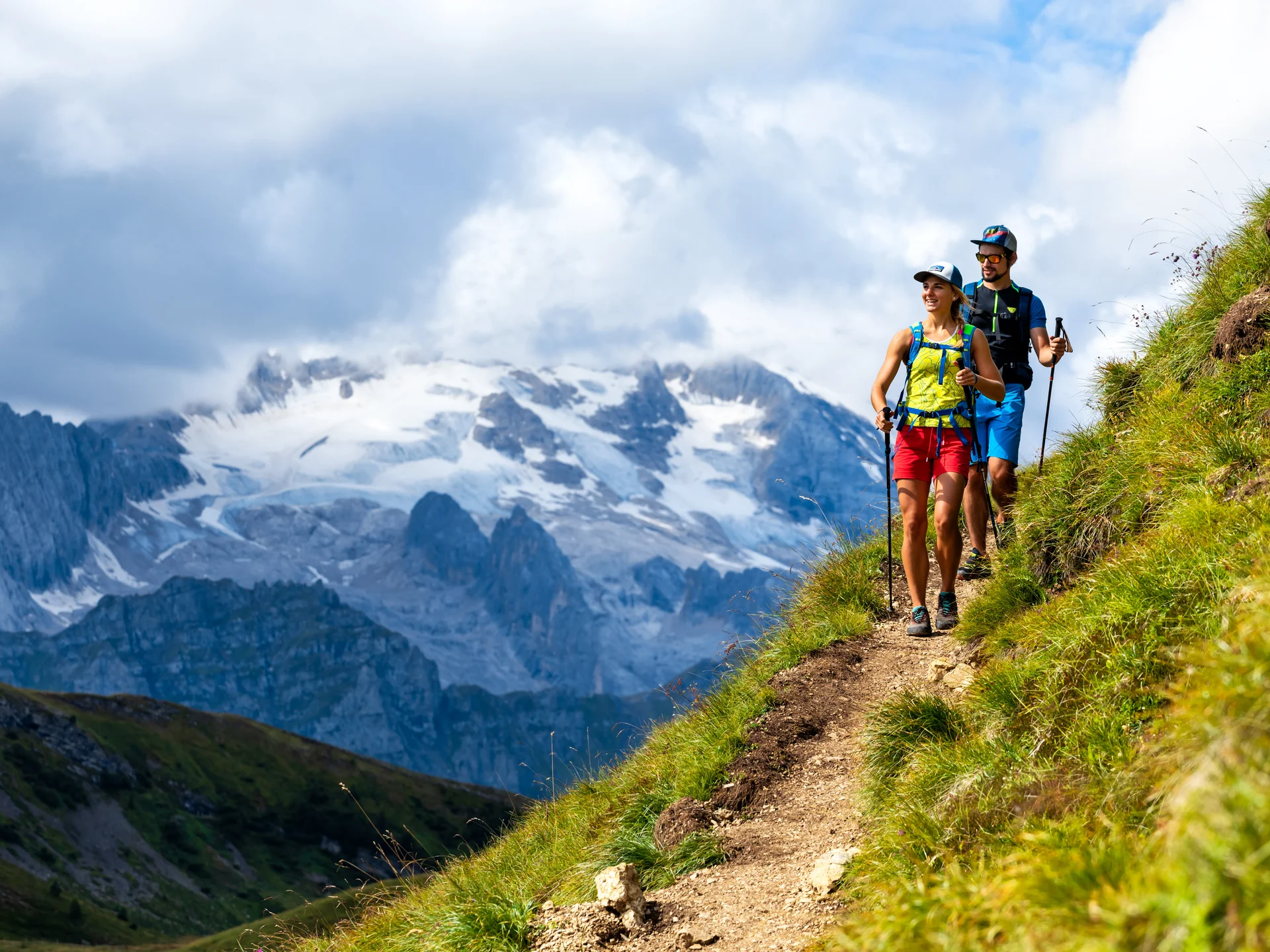 Two hikers on mountain trail with snowy mountains in background