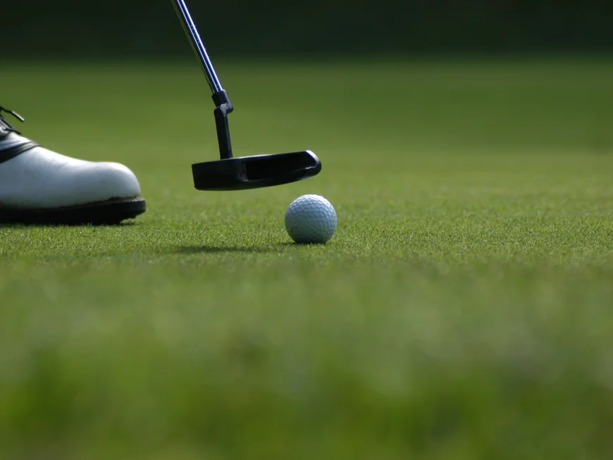 Golf shoe and putter hitting a golf ball on the green grass